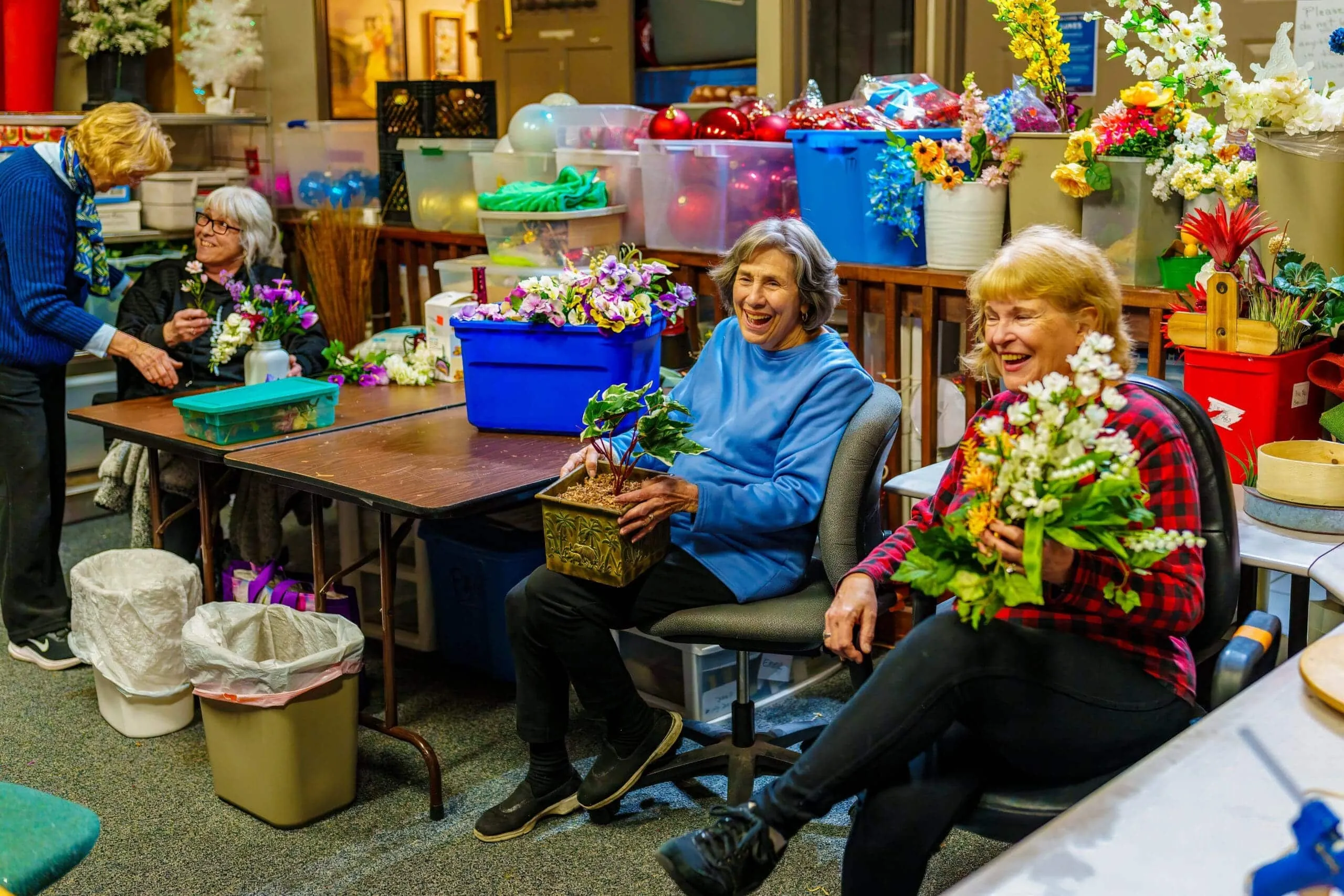 Volunteers arrange artificial flowers at tables in a colorful room filled with craft supplies and decorations.
