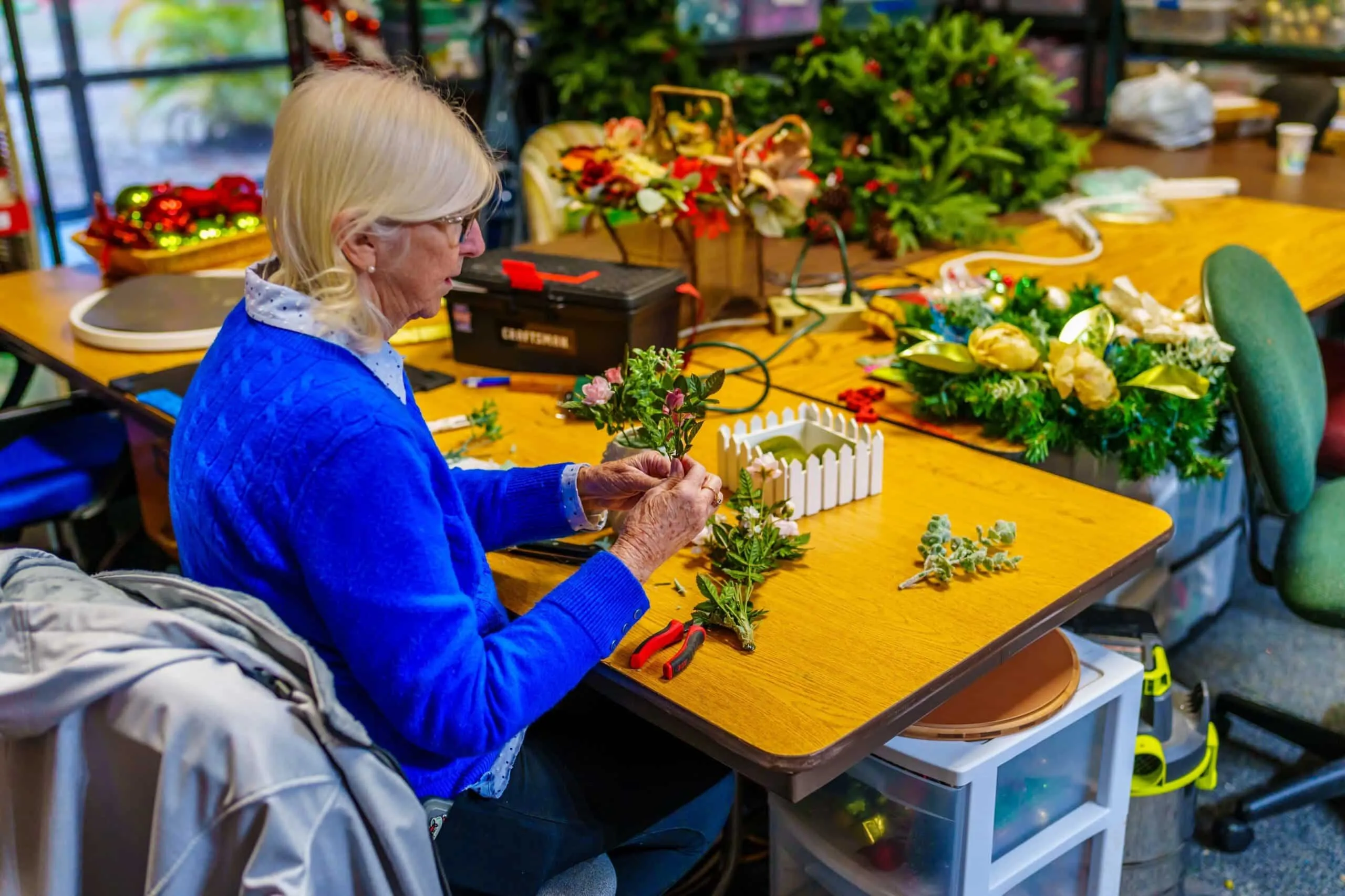 A woman assembles floral decorations at a table filled with craft supplies and greenery.