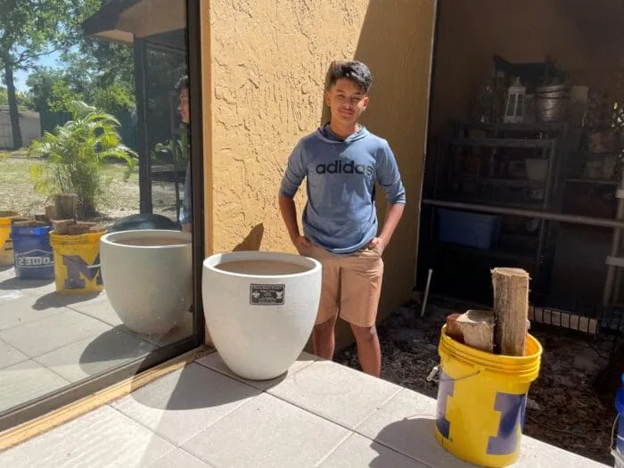 A young person stands next to a large white planter and a yellow bucket filled with logs outside a building.