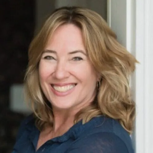 A woman with blonde hair smiles while leaning against a doorway, wearing a blue top.