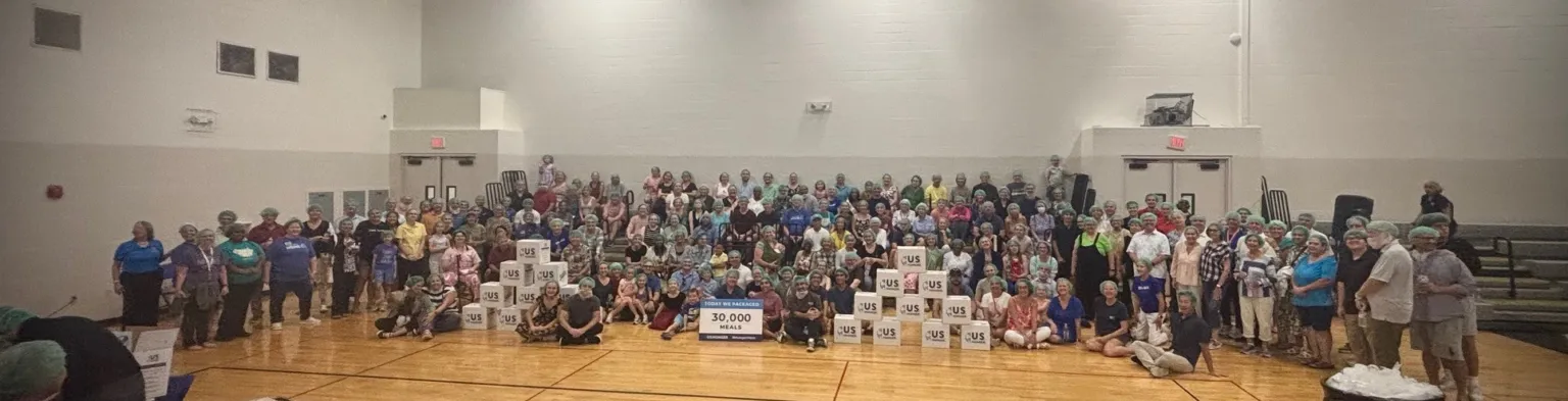 A large group of people stands together in a gymnasium, celebrating the packing of 30,000 meals.
