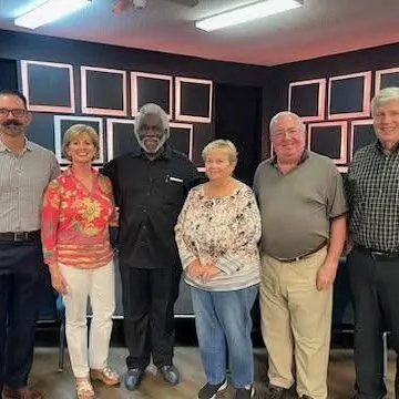 Six people stand together, smiling in front of a dark wall with framed pictures.