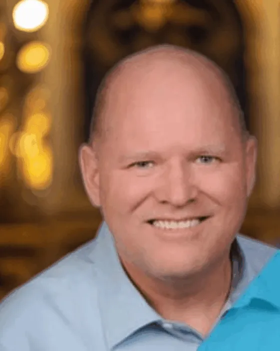 A man with a bald head and a blue shirt smiles in front of a softly lit background.