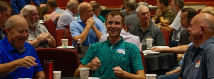 Men are gathered around tables, enjoying breakfast and engaging in conversation at a social event.