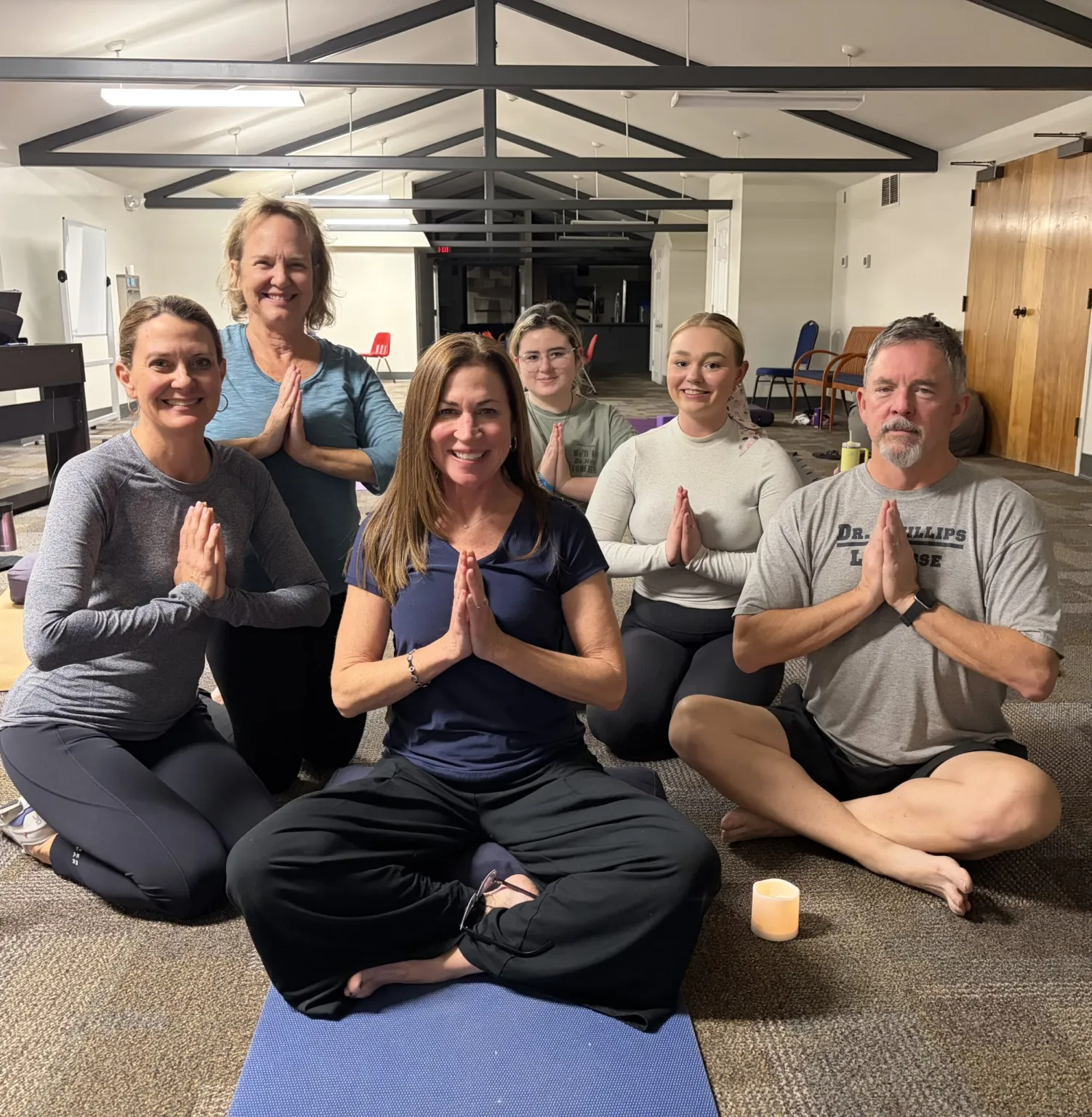 Participants are seated in a yoga class, practicing a prayer pose with hands together.