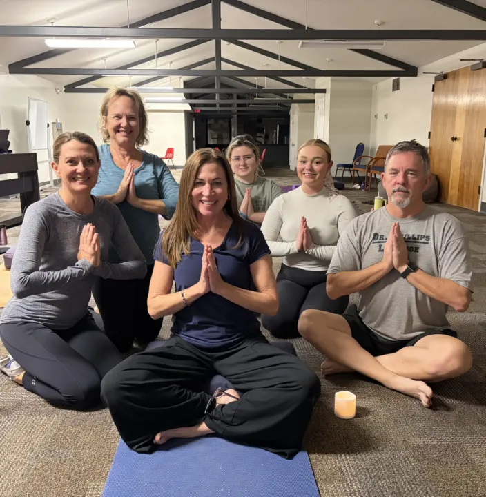 Participants are seated in a yoga class, practicing a prayer pose with hands together.