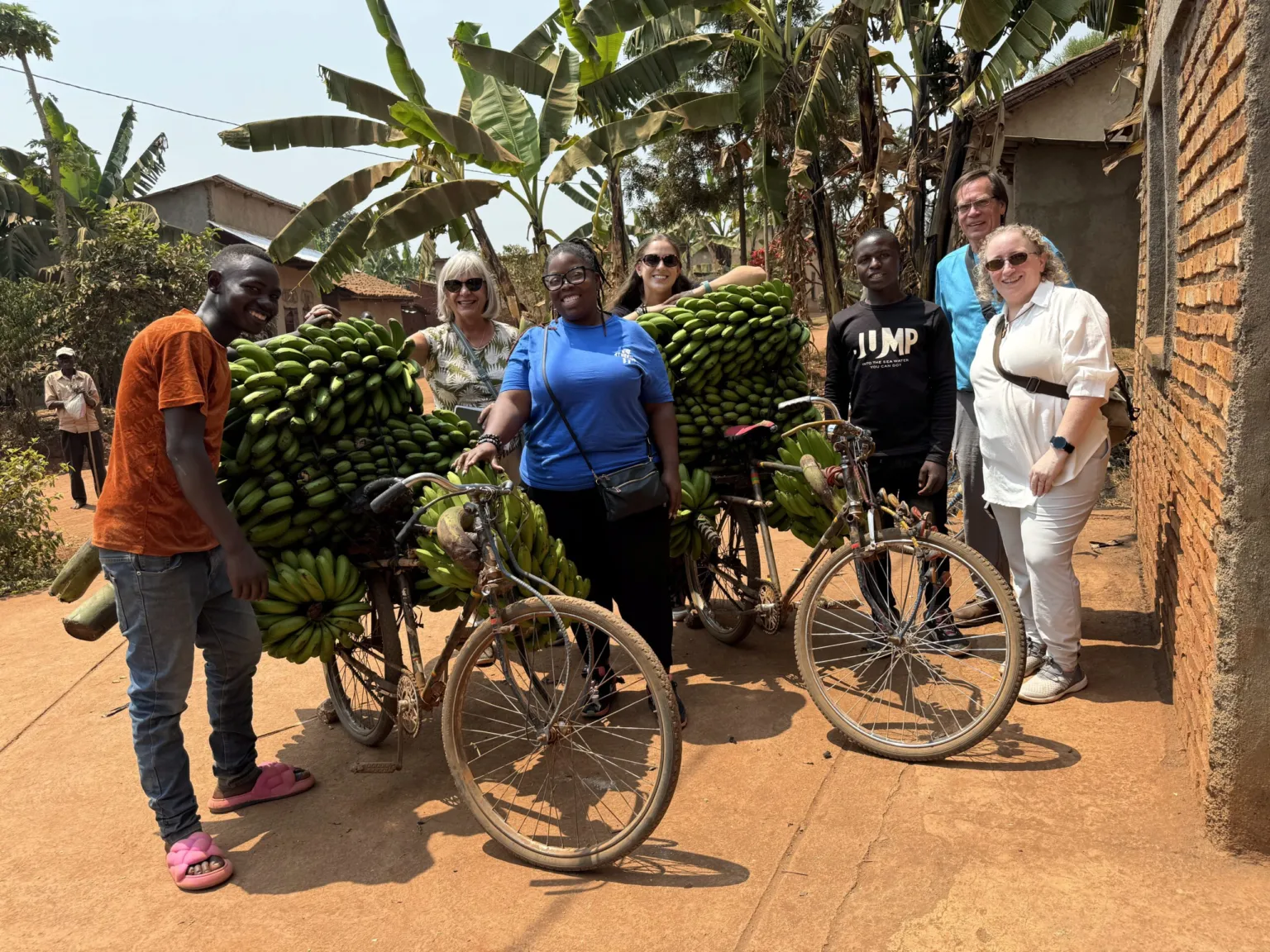 Group of people pose with bicycles loaded with bananas in a rural setting surrounded by greenery and buildings.