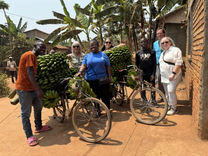 Group of people pose with bicycles loaded with bananas in a rural setting surrounded by greenery and buildings.