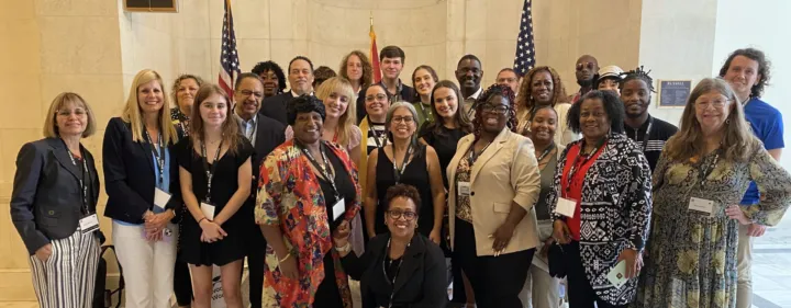A diverse group of people poses together in a hall with American flags, smiling and wearing name tags.