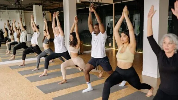 Participants are practicing yoga in a studio, holding a pose with arms raised.