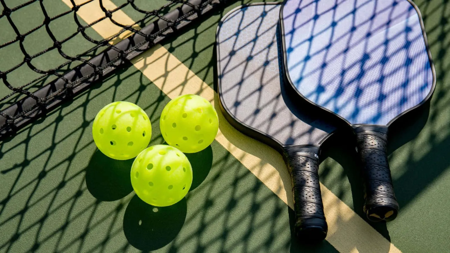 Pickleball paddles and bright yellow balls are placed on a green court beside a net.