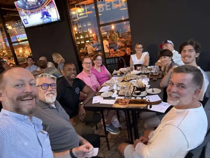 A group of people smiles at the camera while sitting around a table with food and drinks in a restaurant.