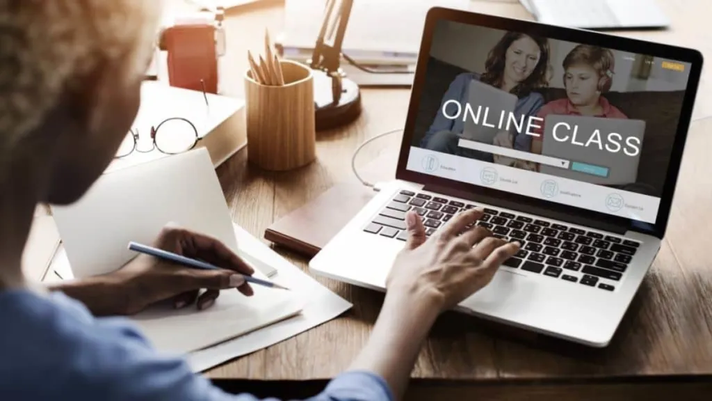 A person writes in a notebook while using a laptop displaying an "ONLINE CLASS" webpage.
