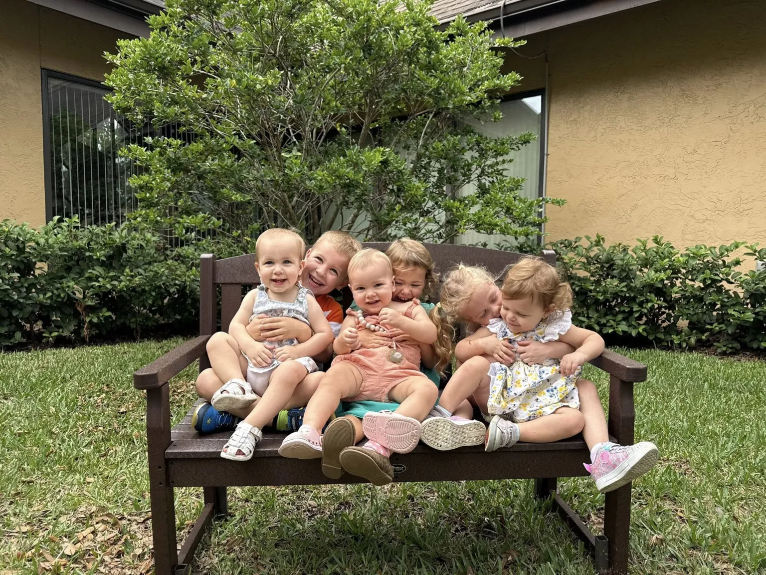 Children sit closely together on a bench outdoors, smiling and hugging each other in a grassy area.