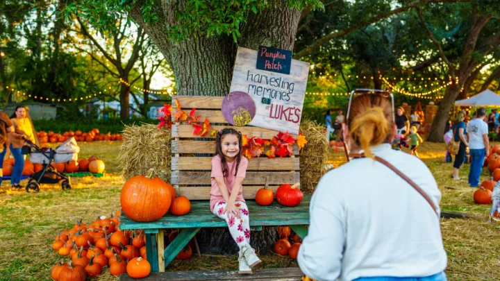 A girl sits on a bench in a pumpkin patch, surrounded by pumpkins and people enjoying the festive atmosphere.