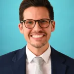 A man in a suit and tie smiles against a light blue background.