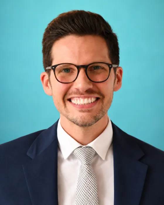 A man in a suit and tie smiles against a light blue background.