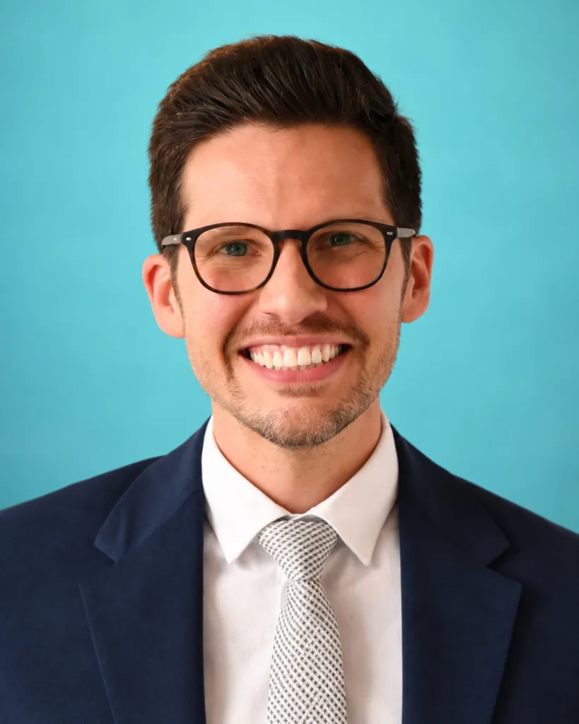 A man in a suit and tie smiles against a light blue background.