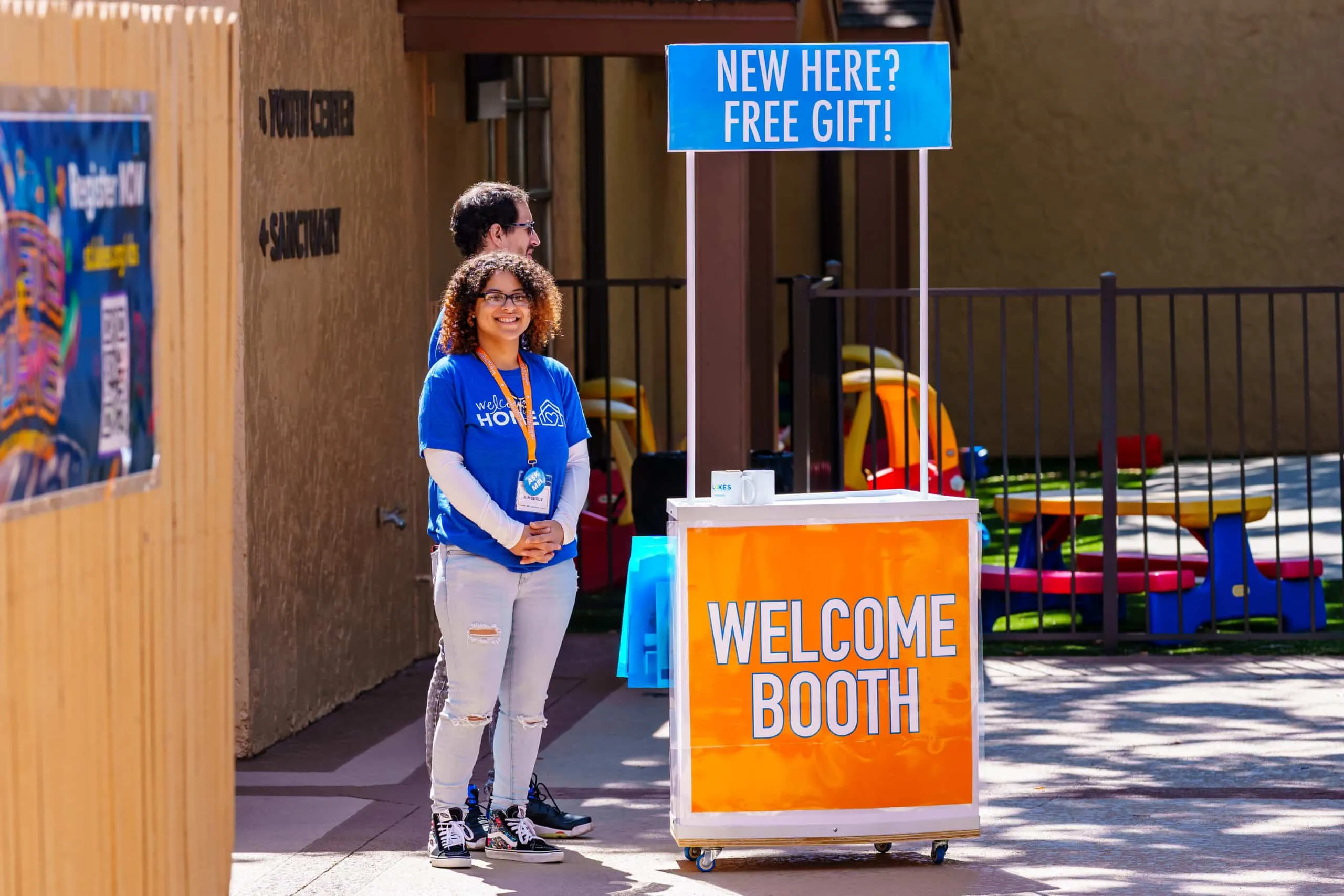 A welcome booth with a smiling attendant offers a free gift to newcomers outside a church.
