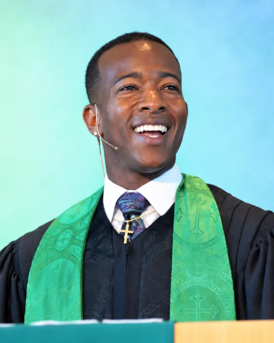 A smiling pastor wearing a green stole delivers a message from behind a podium.
