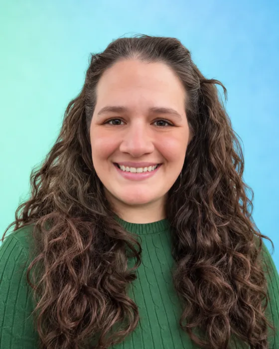 A person with curly hair smiles while wearing a green sweater against a blue and green background.