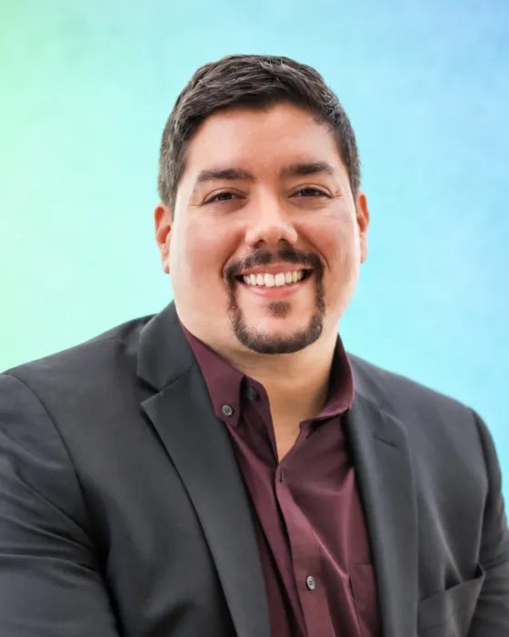 A man in a black suit smiles confidently against a soft blue and green background.