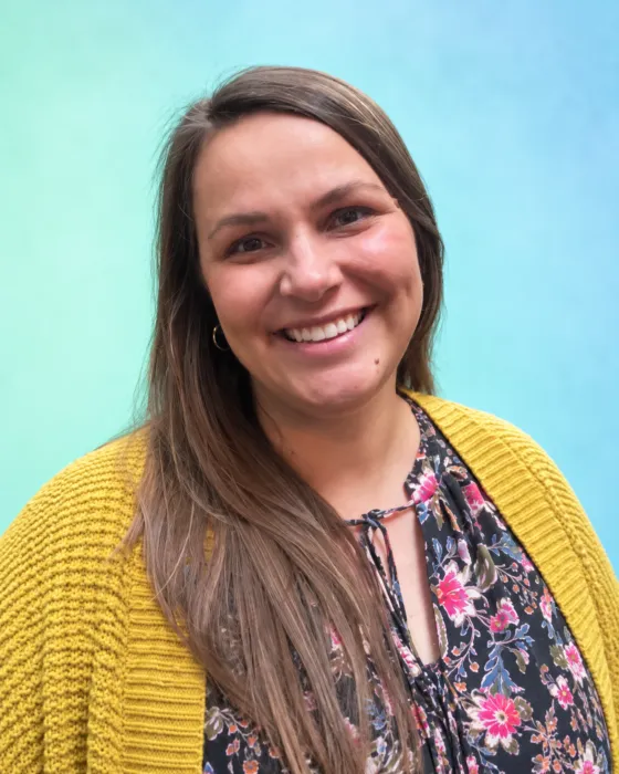 A woman with long hair smiles while wearing a floral top and a yellow cardigan against a blue-green background.