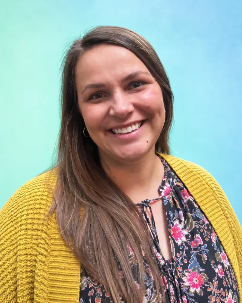 A woman with long hair smiles while wearing a floral top and a yellow cardigan against a blue-green background.