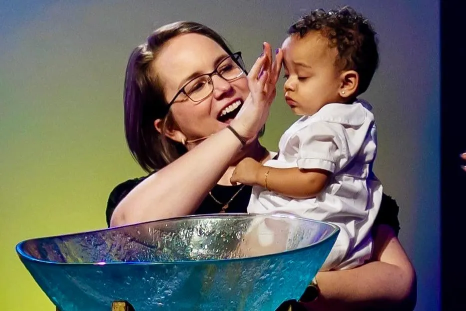 A woman holds a baby near a baptismal font, smiling and interacting with the child during a church ceremony.