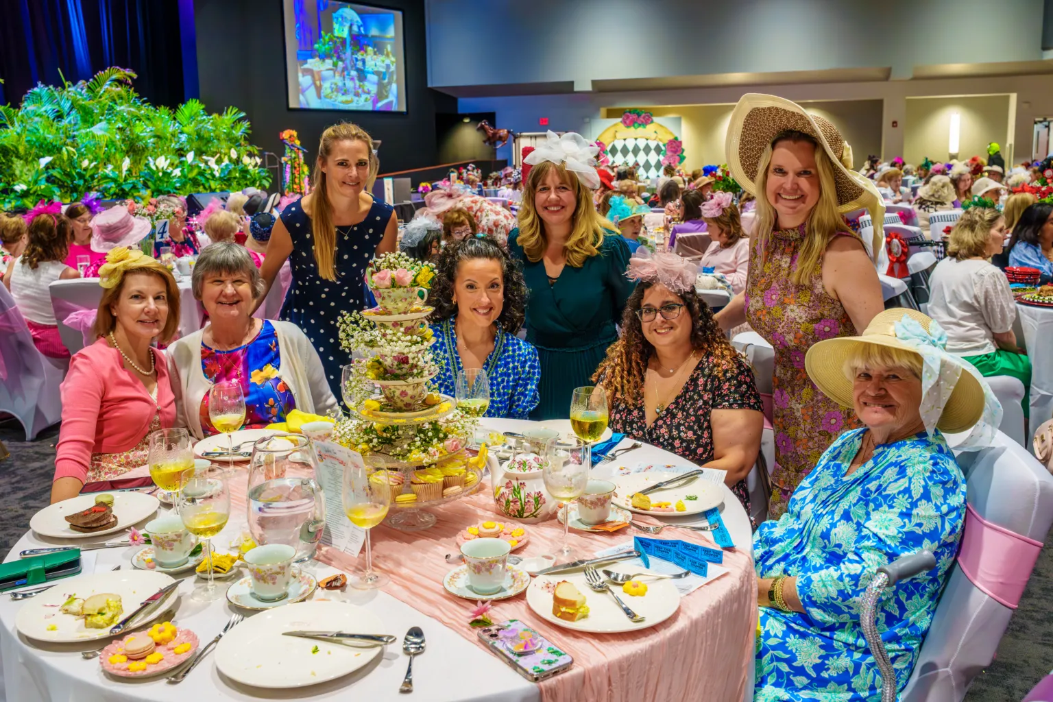 A group of women in hats pose around a table with tea and desserts during a festive high tea event.