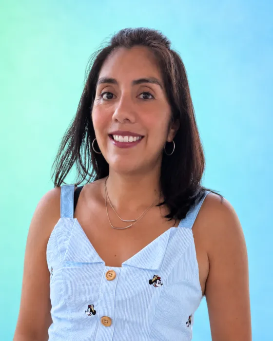 A woman smiles while wearing a light blue dress with buttons and embroidered designs, set against a gradient background.
